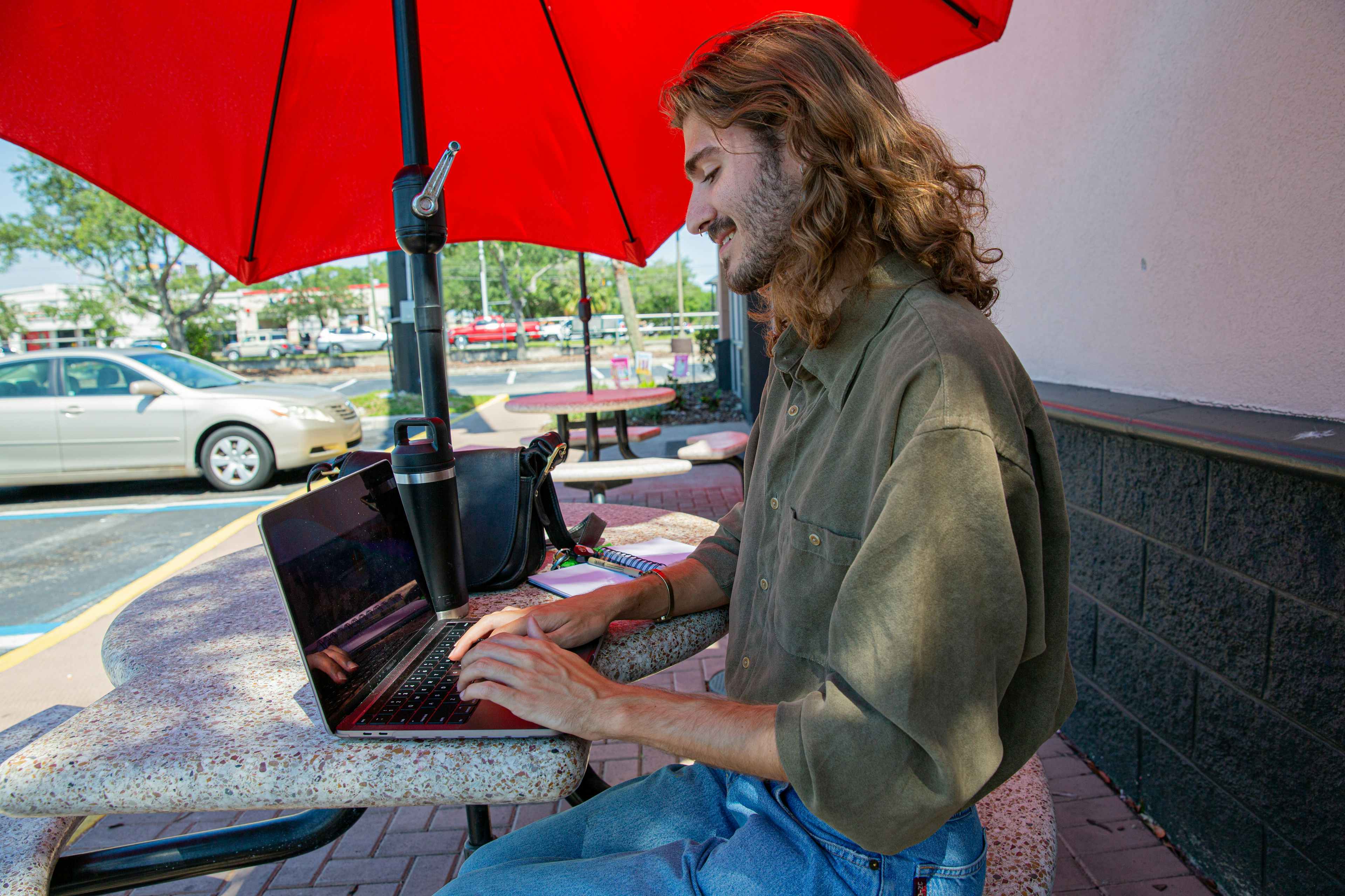 Student typing on a laptop outside, sitting on a bench underneath a red awning
