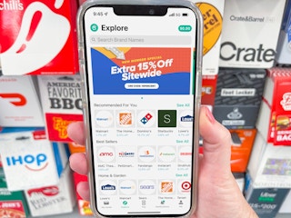 A person holding a cell phone with the Raise app in front of a wall display of physical gift cards.