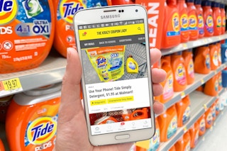 Krazy coupon lady app displaying a deal on laundry detergent, in front of Tide on the shelves of the detergent aisle inside Walmart.