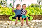 Lifestyle image of a green flying saucer swing on a tree branch over the sand with two small kids sitting on it