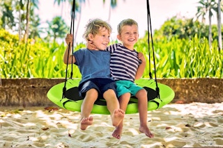 Lifestyle image of a green flying saucer swing on a tree branch over the sand with two small kids sitting on it