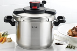A stainless steel T-Fal Clipso Pressure cooker sits on a counter between a cutting board and a plate of food. It has secure-locking 1-handed