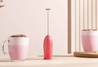 a pink milk frother on a natural wood table with two cups of coffee