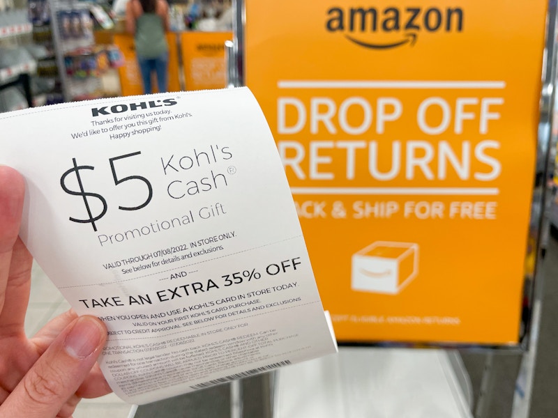 A person's hand holding a $5 Kohl's Cash coupon on a receipt for returning an Amazon package next to the Amazon drop off returns sign ins...