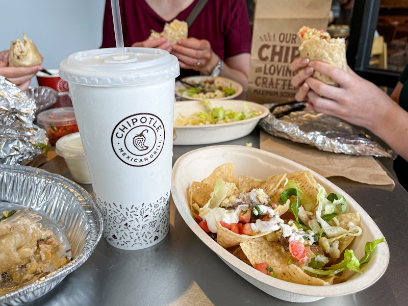 Three people holding burittos at a table filled with food. A drink cup is in the for front clearly displaying the Chipotle logo.