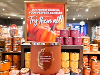 A person's hand holding up a fall 3-wick candle in front of a display of fall candles in Bath and Body Works.