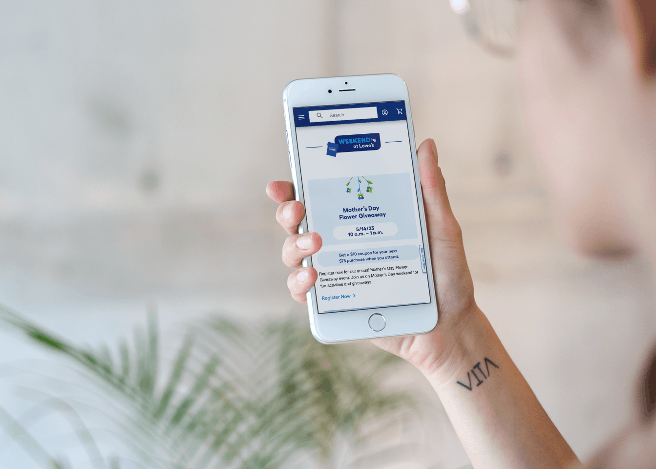 A woman holding up her phone and looking at the Lowe's event page online, showing a $10 coupon is available if you attend the Mother's Da...