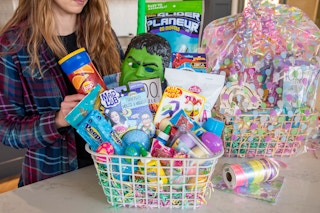 A woman assembling an Easter basket with a second one, wrapped in an Easter themed basket bag in the background.