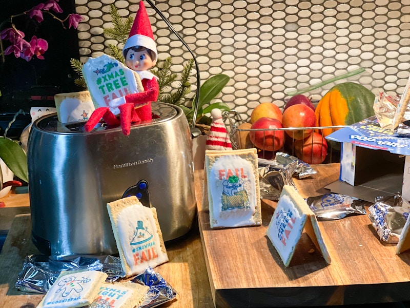 an elf on the shelf doll sitting on a toaster playing with poptarts 