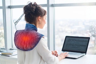Lifestyle image of a woman wearing a weighted heating pad over shoulders while using a computer