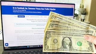 Dollar bills being held in front of laptop with facebook settlement on screen