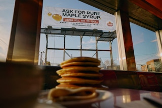 A stack of pancakes in the foreground with the "Bill the Maple" billboard behind it.