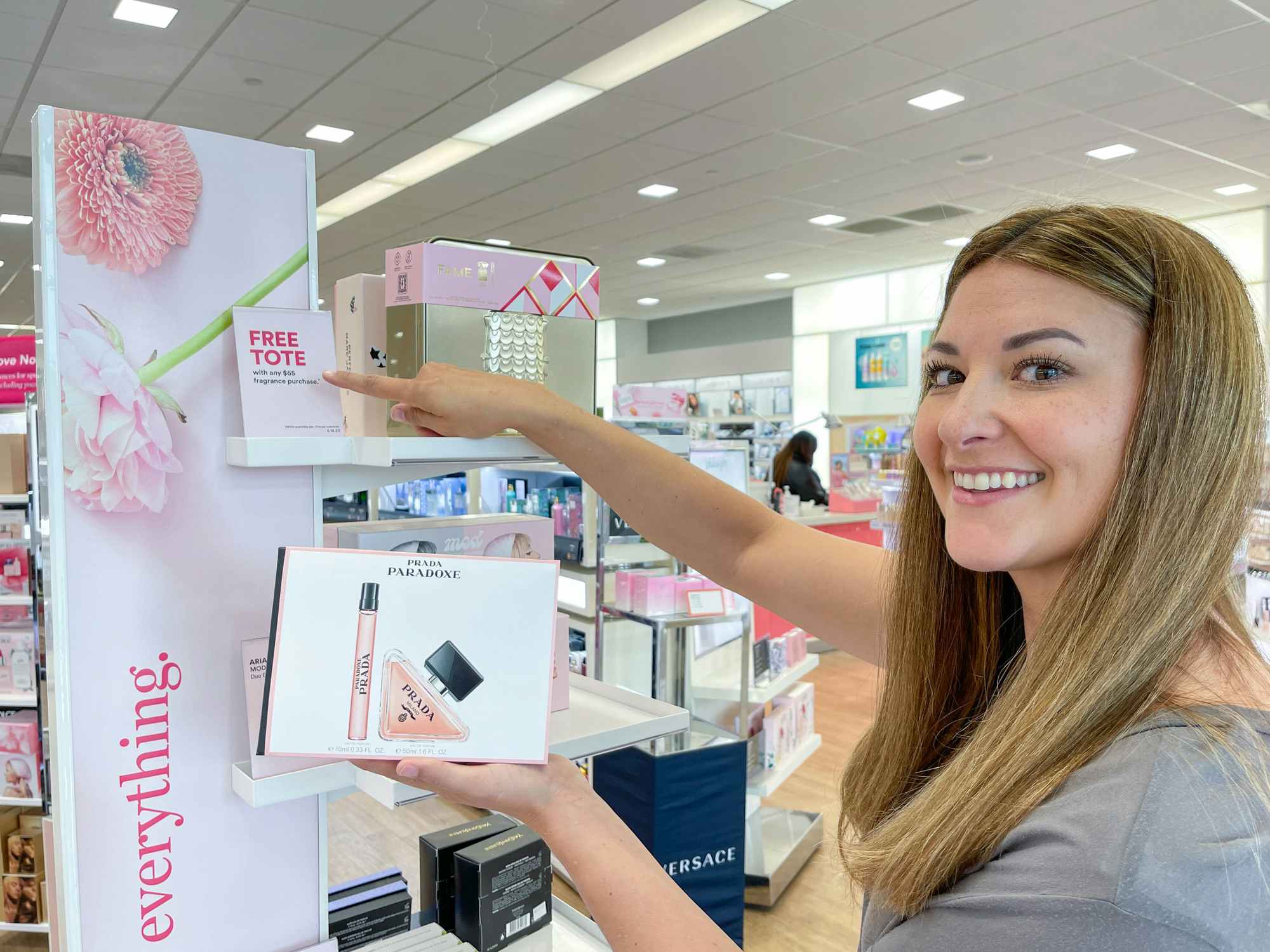 A person holding a fragrance product and pointing to a sign advertising a free tote with purchase