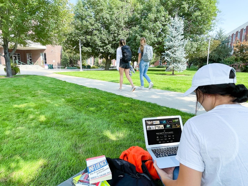 A college student sitting in grass on campus and watching Apple TV on laptop with two more students walking by on the sidewalk in the ...