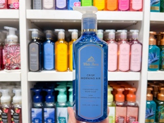 A hand soap held out by hand in front of a shelf with other hand soaps on it.