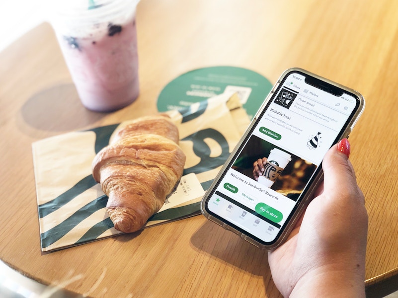 A person holding a smartphone with the Starbucks app open above a table with a croissant and drink sitting on it.