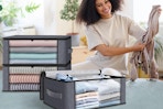 A young woman sits on her bed folding clothes and putting them into 3 under bed storage bags with clear windows and handles.