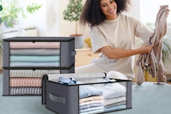 A young woman sits on her bed folding clothes and putting them into 3 under bed storage bags with clear windows and handles.