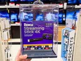 A Roku Streaming Stick 4k held out by hand in front of aisle.