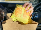 A person pulling a wrapped McDonald's cheeseburger out of a McDonald's bag while sitting in their car.