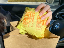 A person pulling a wrapped McDonald's cheeseburger out of a McDonald's bag while sitting in their car.