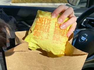 A person pulling a wrapped McDonald's cheeseburger out of a McDonald's bag while sitting in their car.