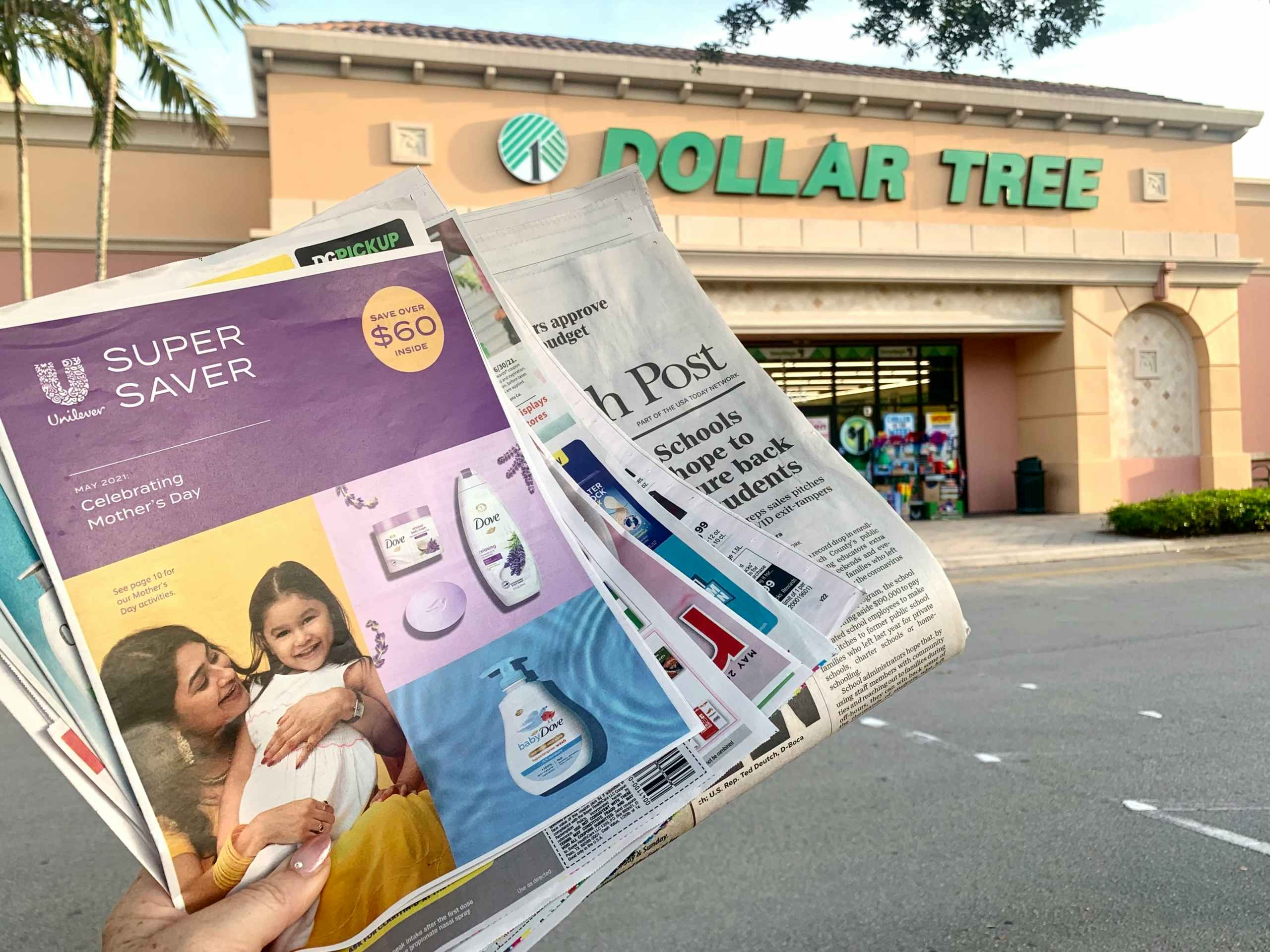 A person’s hand holding a stack of coupon leaflets and a newspaper in front of a Dollar Tree storefront.