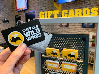 Someone holding two Buffalo Wild Wings gift cards in front of the Gift Card stand inside Buffalo Wild Wings.