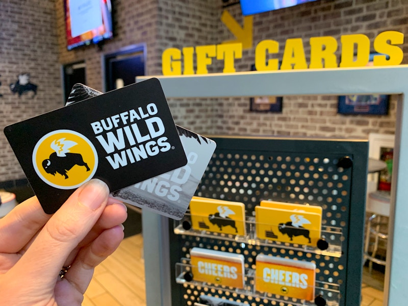 Someone holding two Buffalo Wild Wings gift cards in front of the Gift Card stand inside Buffalo Wild Wings.