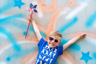 A child with American flag sunglasses laying down on a patriotic firework photo backdrop drawn on concrete with spray chalk.