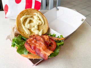 A bacon lettuce tomato sandwich open in a cardboard takeout box from chick-fil-a
