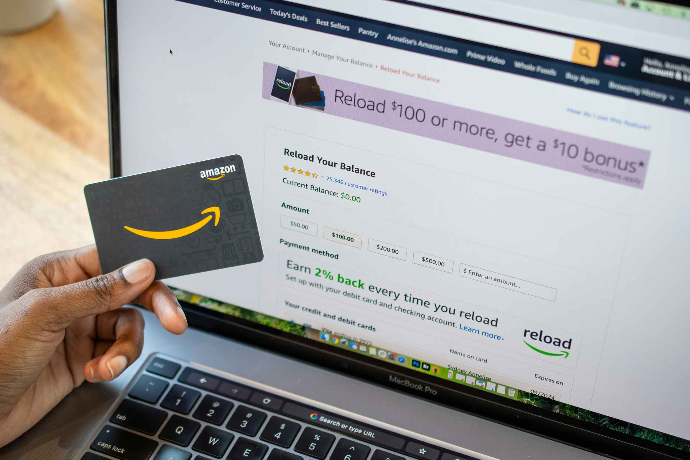 A person's hand holding an Amazon gift card in front of a laptop screen displaying the Amazon gift card reload webpage.