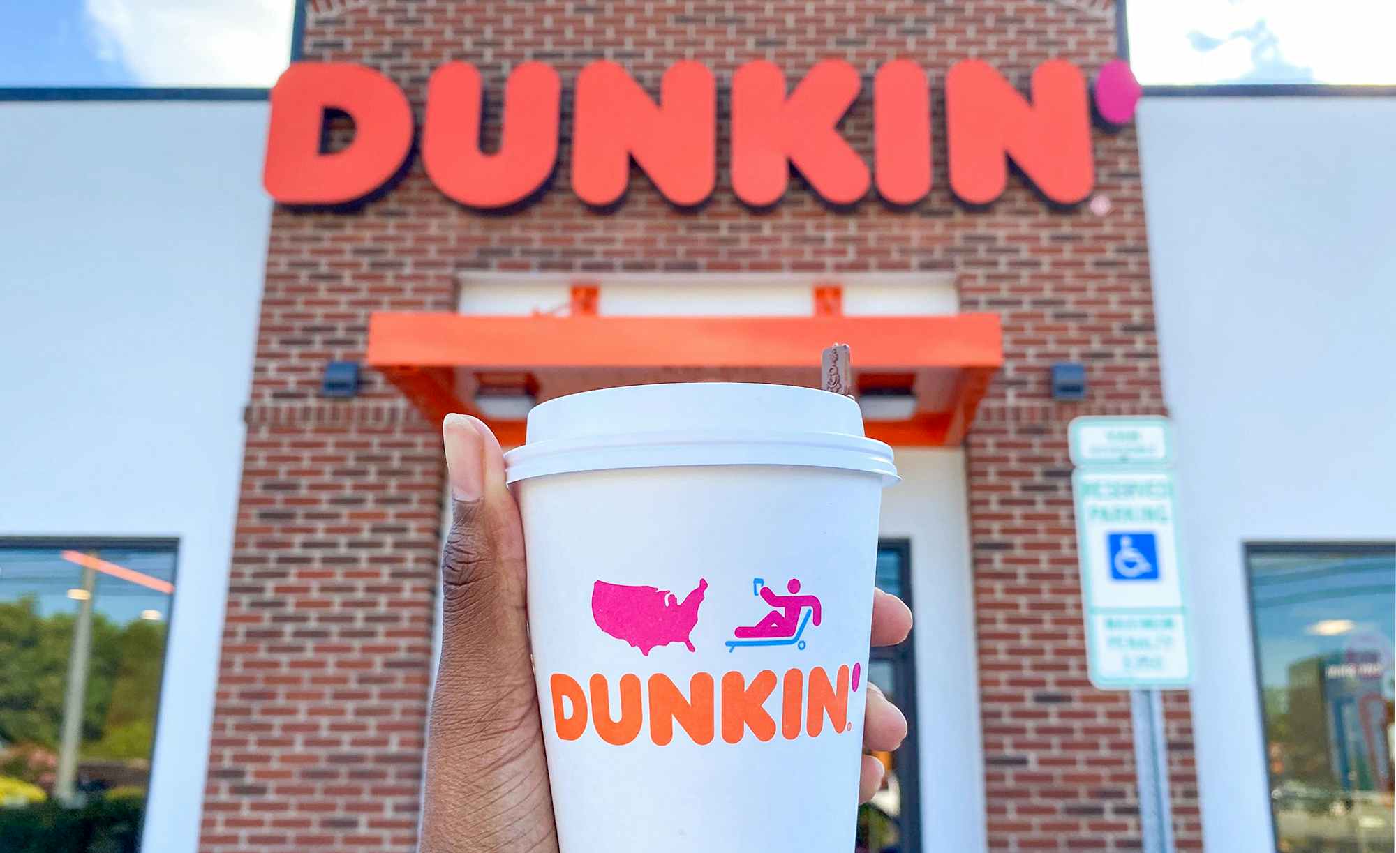 person holding a hot cup of dunkin donuts coffee in front of restaurant entrance