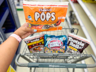 hand holding bag of Tootsie Pops with two bags of Tootsie Rolls and Double Bubblegum inside of shopping cart in background