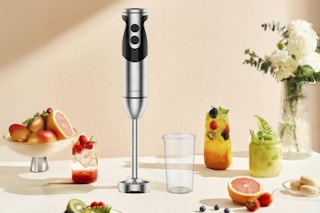 A hand blender stands on a table among fruit and flowers.