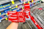 person holding 3 colgate optic white toothpastes over a cvs cart