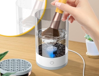 a hand dipping a makeup brush in a electric cleaner on a wooden table