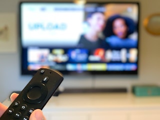 An Amazon fire tv stick remote pointed at a TV with programs out of focus in the background.