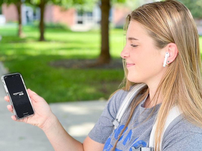 A college student with Airpods in her ears and the Amazon Music app start screen displayed on her phone.