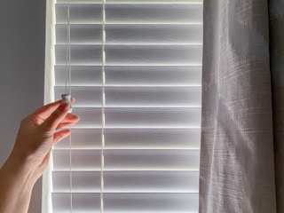 A person's hand closing the blinds of a window where the sun is shining through.