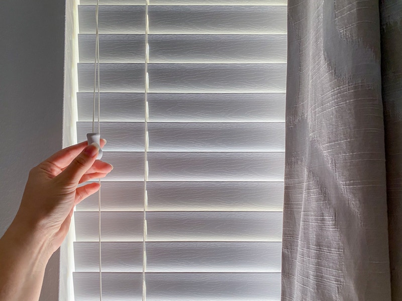 A person's hand closing the blinds of a window where the sun is shining through.