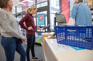 A Walgreens shopping basket on the counter. Two women standing at the checkout with one using the credit card reader to purchase items.