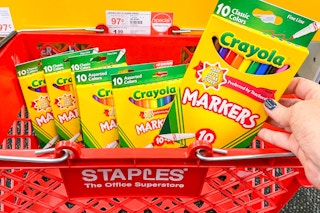 Someone putting some Crayola markers into a Staples shopping basket