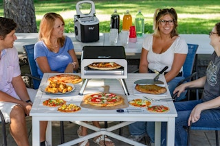 Four people sit around a table that holds dinner and a portable indoor-outdoor electric pizza oven.