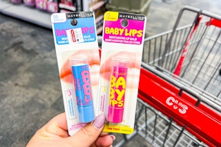 hand holding two Maybelline Baby Lips in front of shopping cart