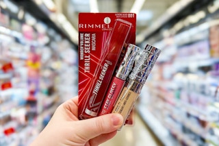 hand holding three rimmel cosmetics in a store aisle