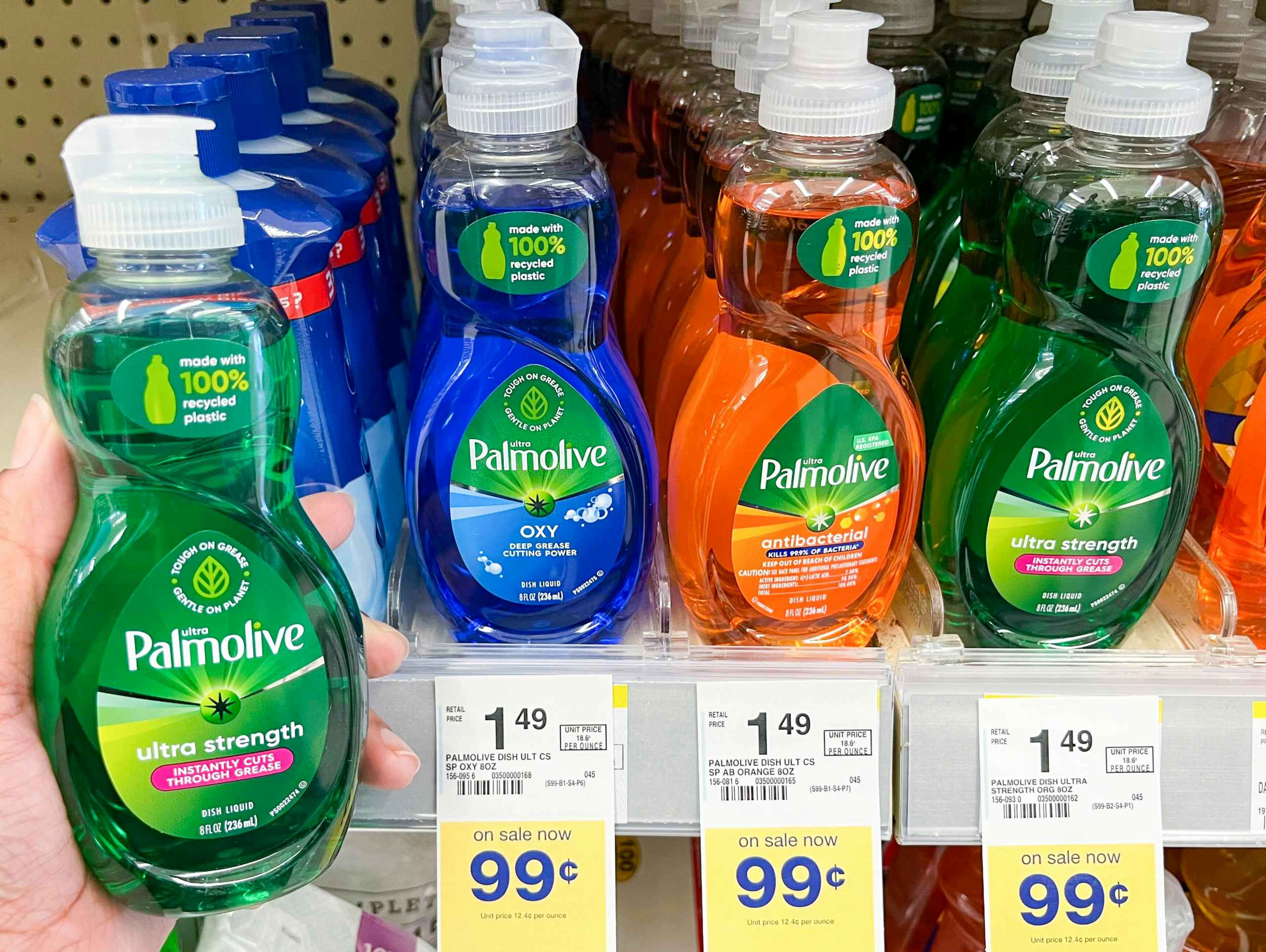 A person's hand holding a bottle of Palmolive Ultra Strength liquid dish soap next to a shelf of different variations of Palmolive liquid...