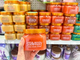 A jar of tree hut strawberry shea sugar scrub held with hand out in front of a store shelf with a variety of tree hut shea sugar scrub ...