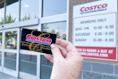 A Costco Membership card being held in front of the Costco store hours sign on the outside of the building.