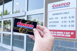 A Costco Membership card being held in front of the Costco store hours sign on the outside of the building.